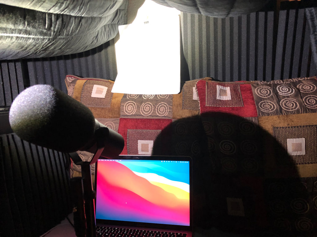 View from inside a sound blanket tent. A MacBook Air and Shure SM7B microphone with pillows and sound panels packed around. A light illuminates the story for narration.