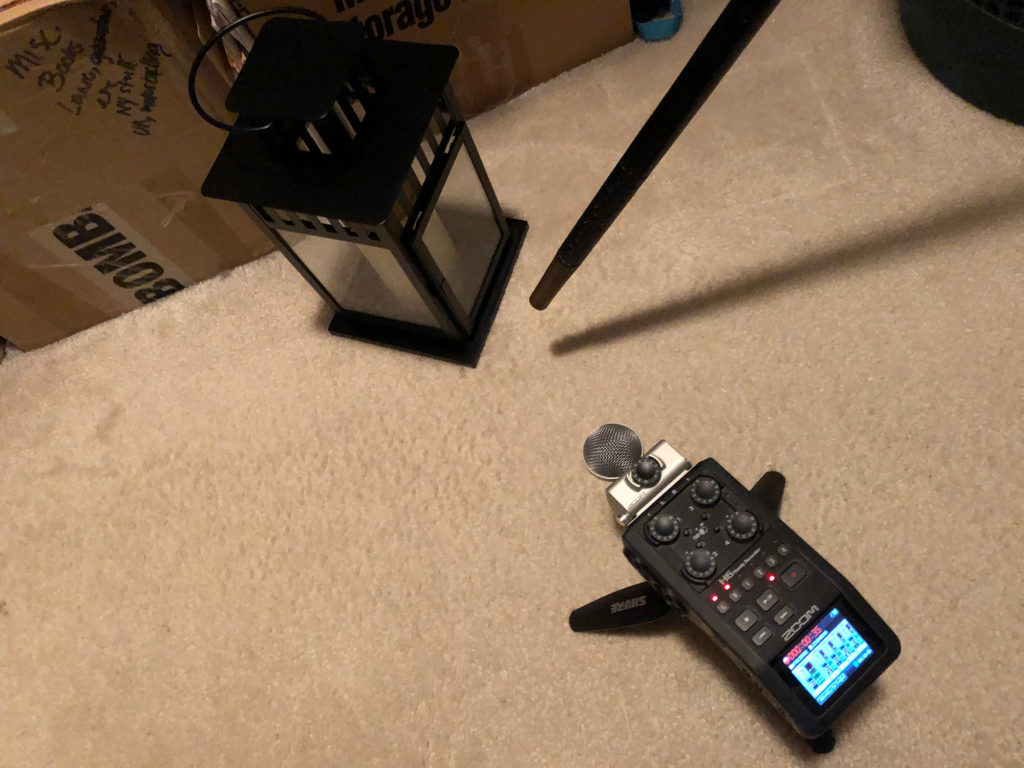 View of a carpeted closet floor. A Zoom H6 portable recorder is on a Shure mini tripod. The bottom of a cane taps a black Ikea candle lantern.