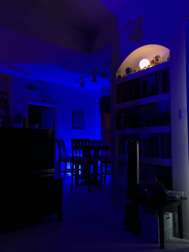 A living room and small dining room flooded in blue LED light. High up on a built-in shelf, a white orb LED glows.